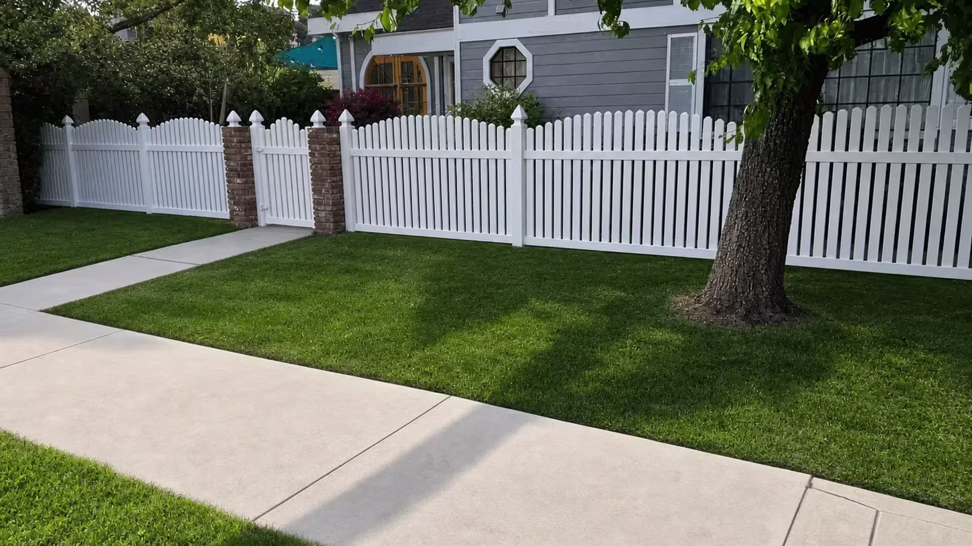 Brick entry columns and a mature tree canopy shelter this White vinyl arched picket fence near the walk
