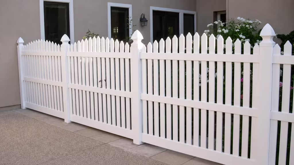 A paved courtyard patio with a wall mounted lantern surrounds this White vinyl arched picket fence