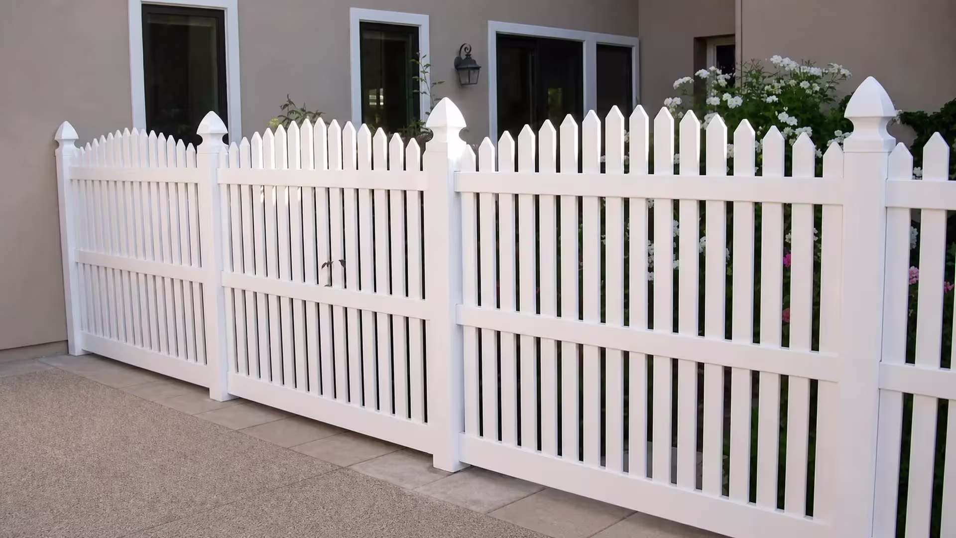 A paved courtyard patio with a wall mounted lantern surrounds this White vinyl arched picket fence