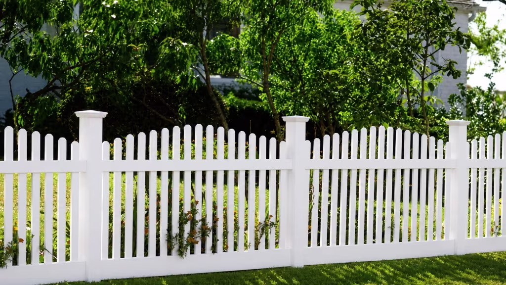 White vinyl arched picket fence with low plants growing through its lower rail along a tree lined lawn