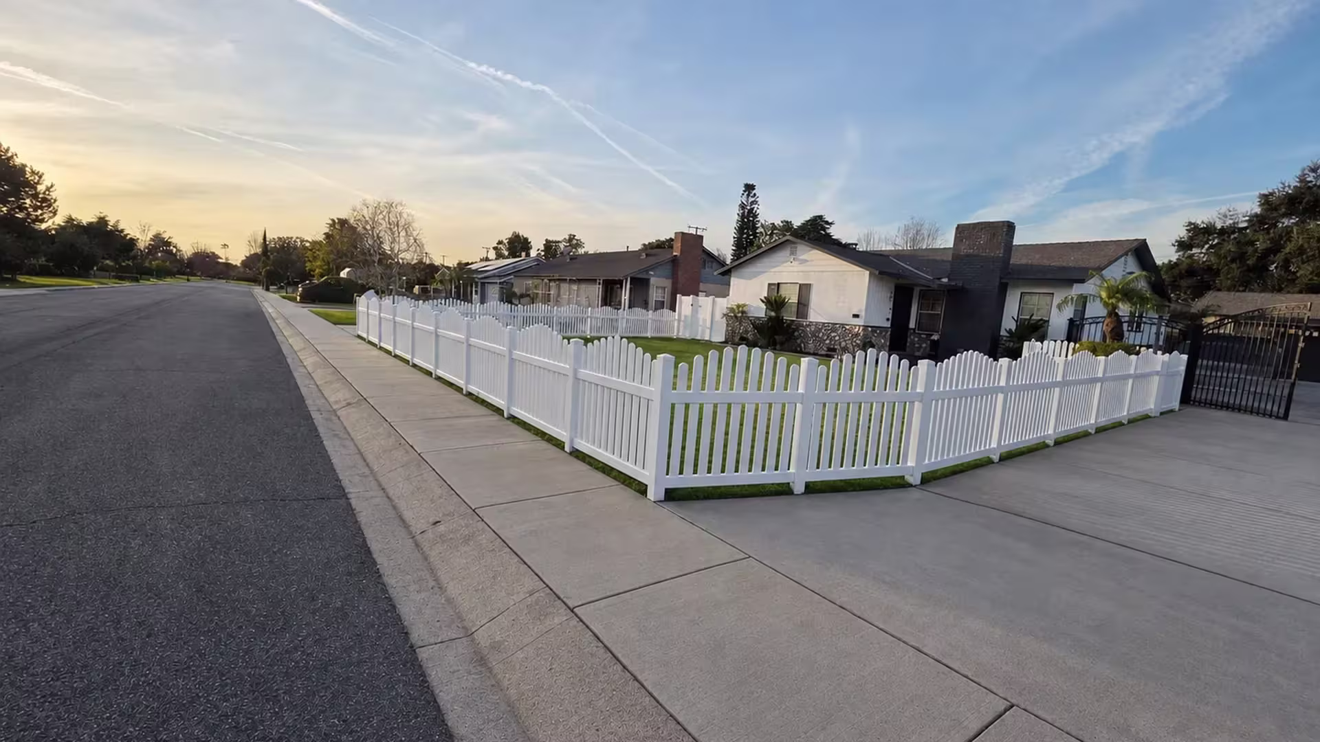 A stone facade home and a separate black metal gate sit beside this White vinyl arched picket fence corner
