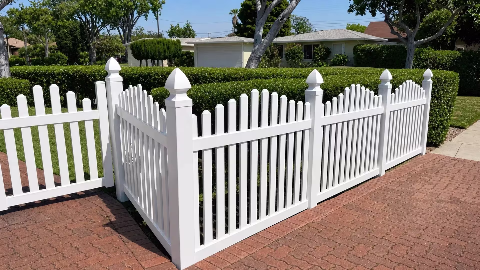 Brick paved driveway edges frame a White vinyl arched picket fence running past palms and clipped hedges