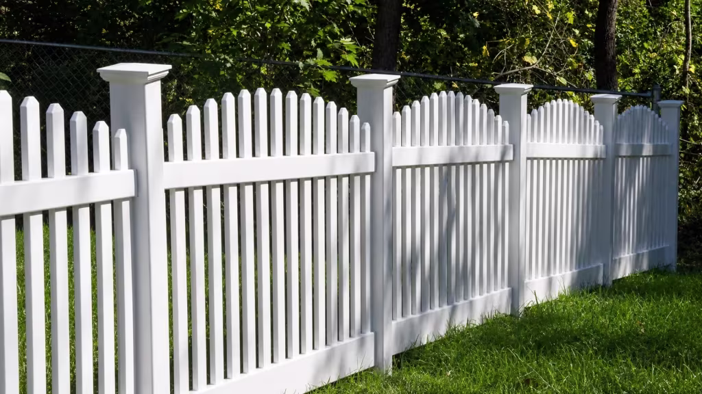Shaded woodland flanks this White vinyl arched picket fence angling diagonally across the yard setting