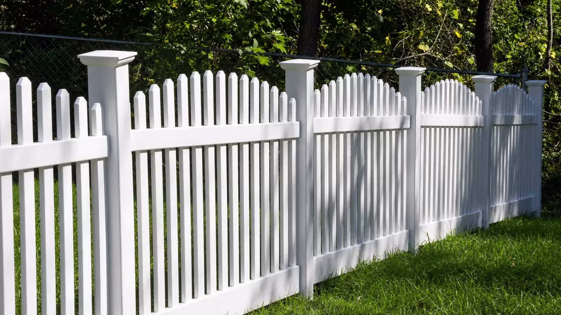 Shaded woodland flanks this White vinyl arched picket fence angling diagonally across the yard setting