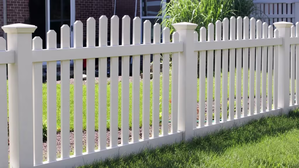 Planting beds sit below a brick house facade fronted by White vinyl arched picket fence