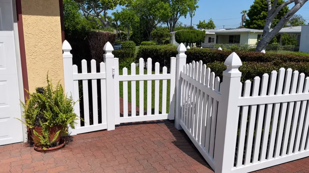 A potted fern and an older chain link fence flank the gated walkway closed by White vinyl arched picket fence