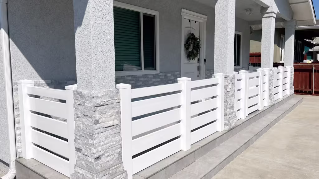 A low porch enclosure of White vinyl fence sits between gray stacked-stone stucco columns