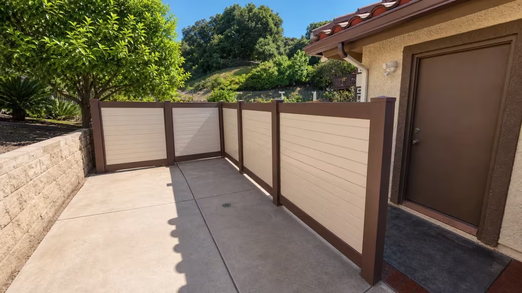 Cream and dark brown two-tone vinyl fence boards run six bays along a hillside stucco yard