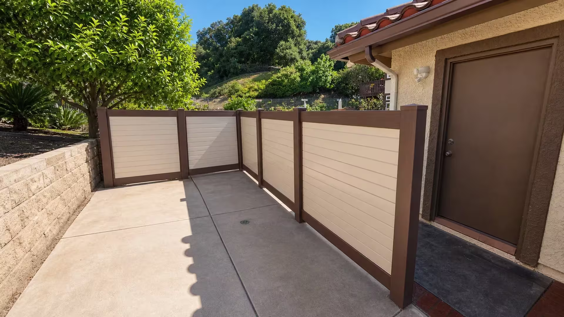 Cream and dark brown two-tone vinyl fence boards run six bays along a hillside stucco yard