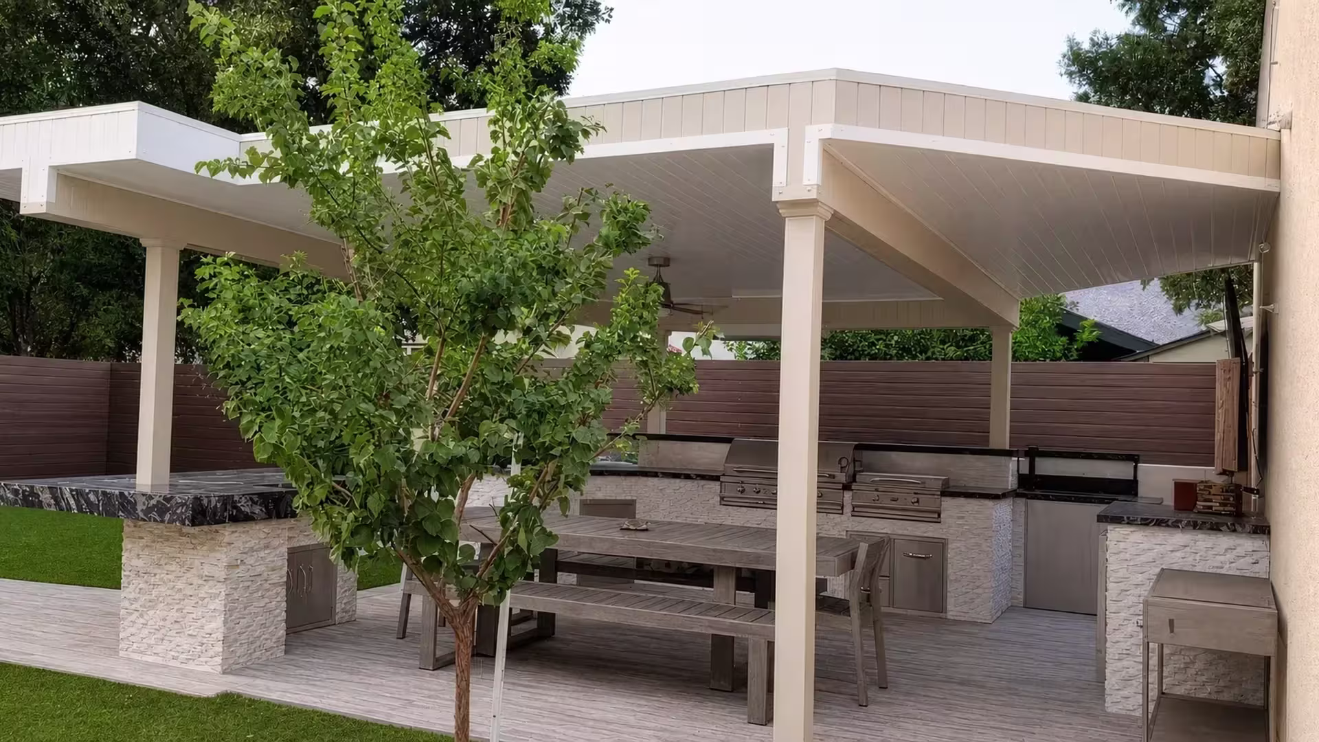 Tan vinyl custom patio cover shading a stone-faced outdoor kitchen and gray plank dining deck