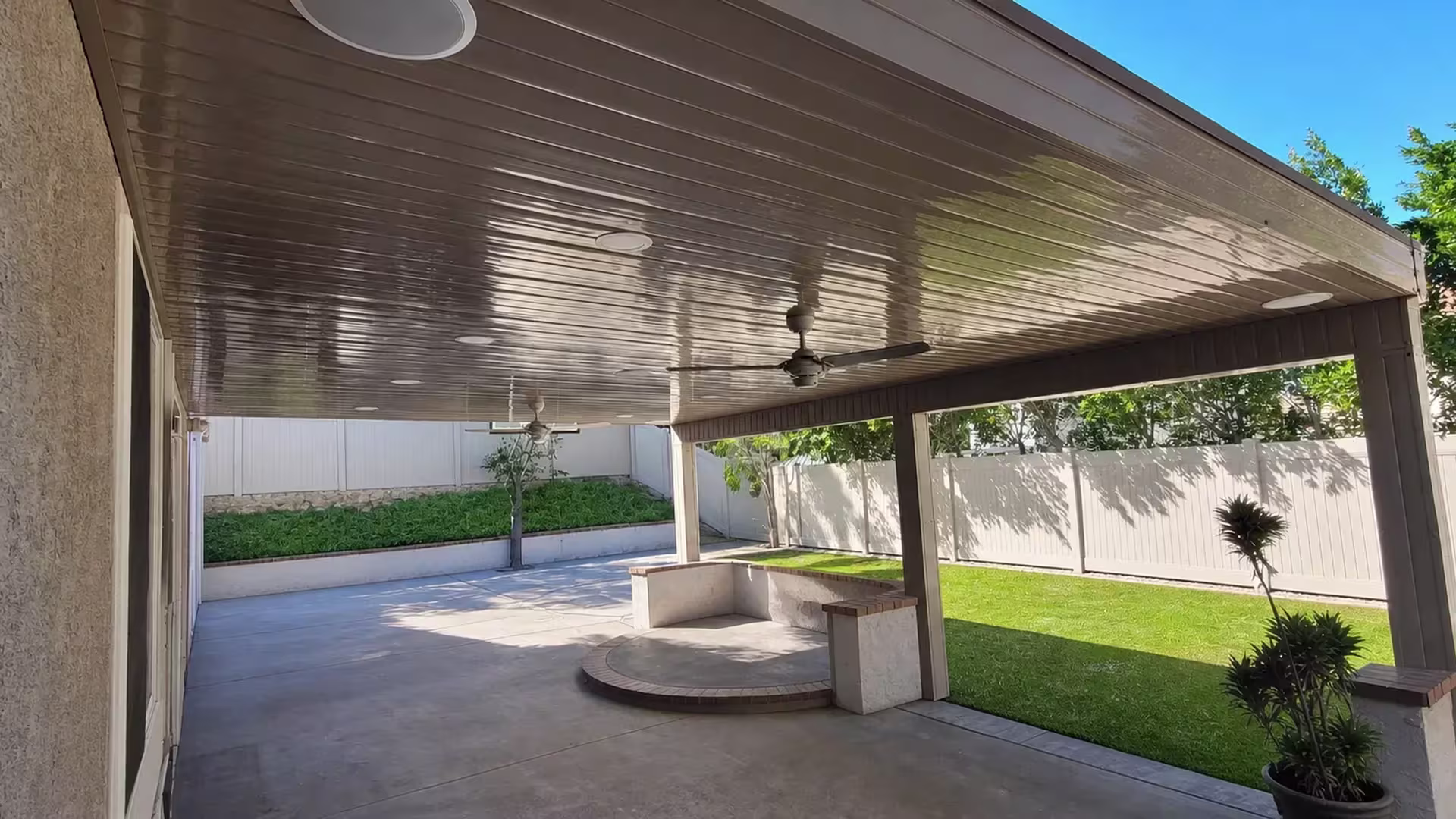 Positioned beside a lawn with curved raised hardscape, this custom patio cover in Tan vinyl fronts a white perimeter fence