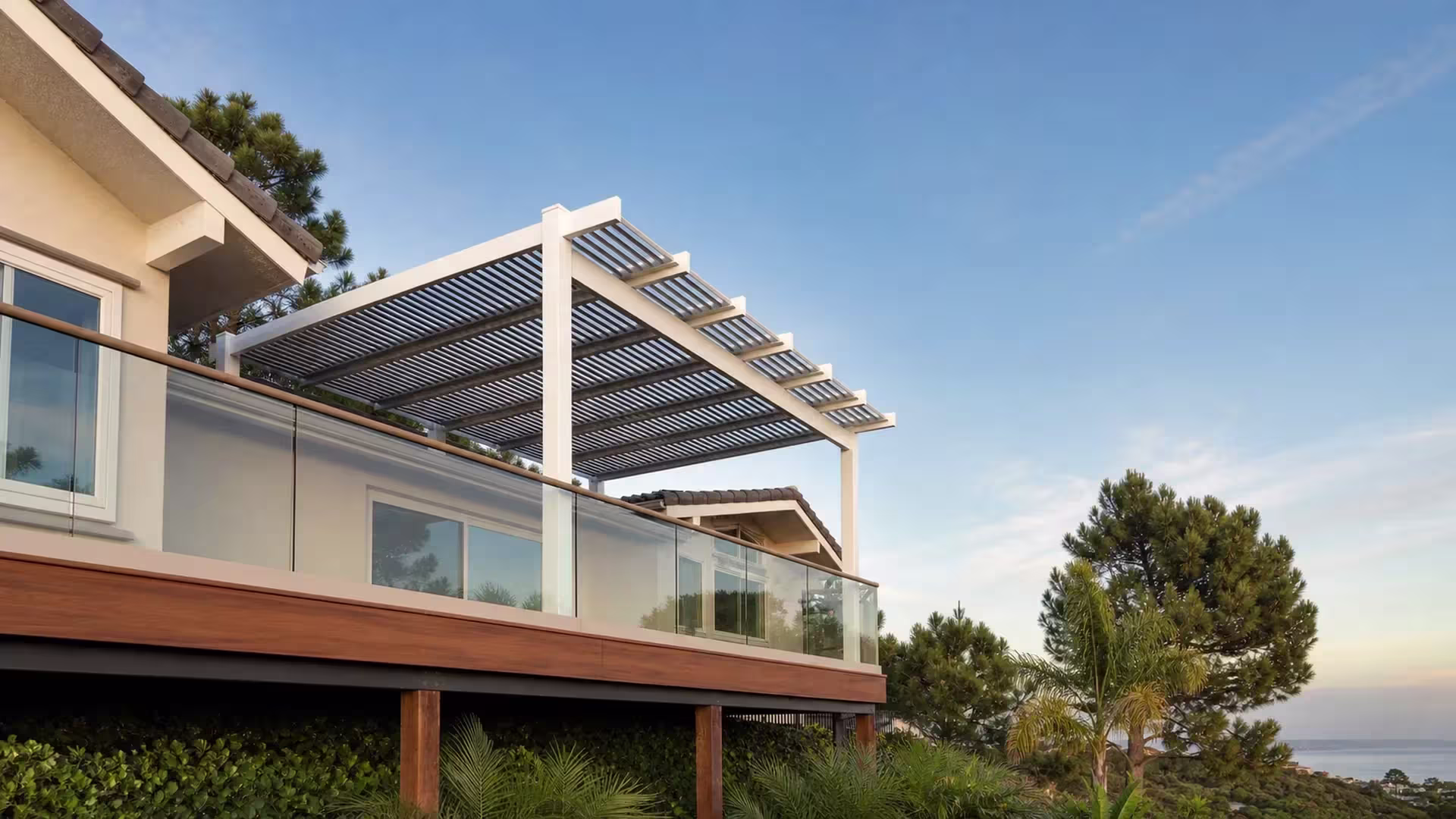 Tiled neighboring rooflines and a glass railing frame the louvered roof of this custom-fabricated White vinyl patio cover