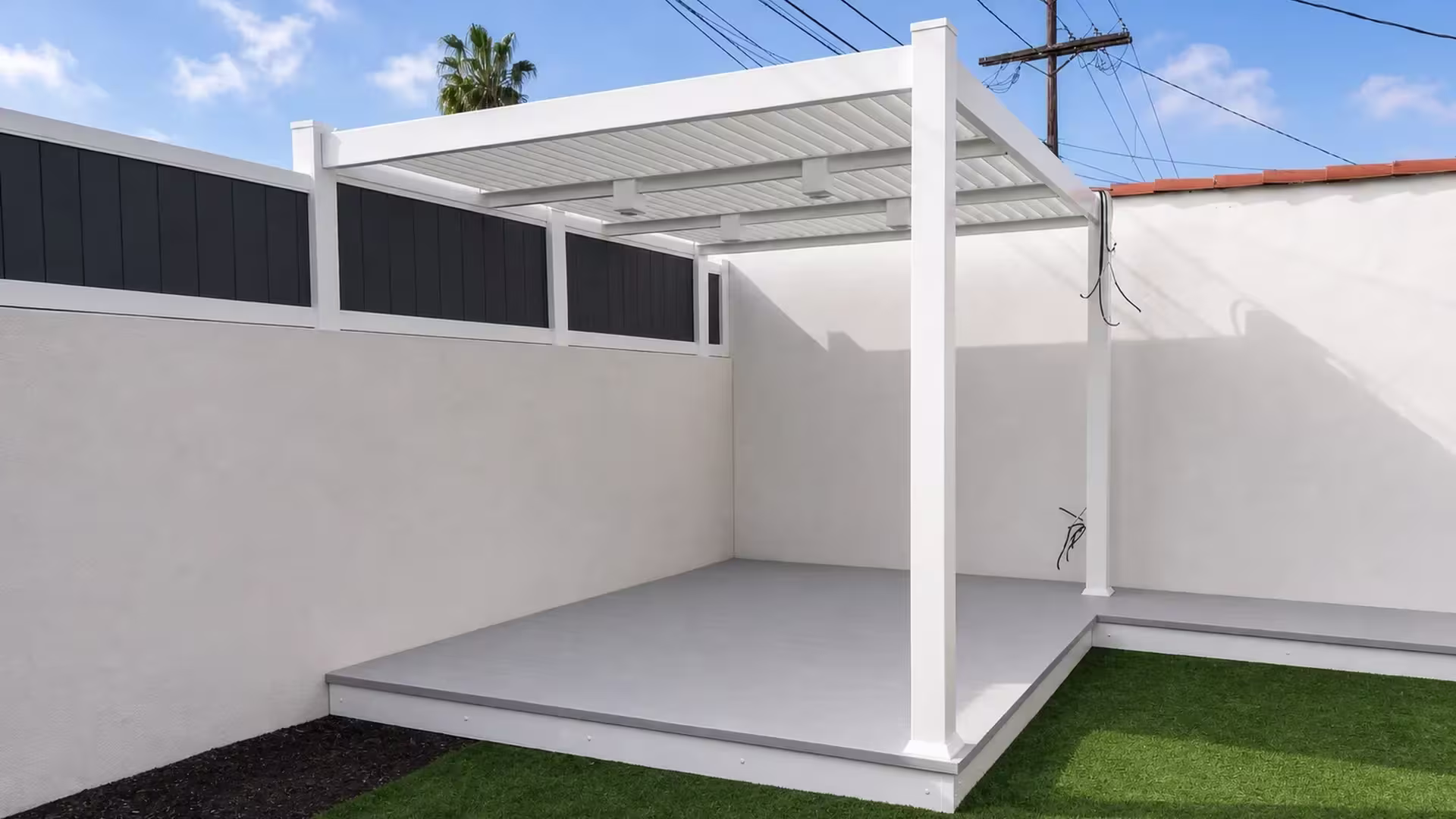 A raised deck bordered by a dark panel fence and palm tree sits beneath a louvered White vinyl custom patio cover
