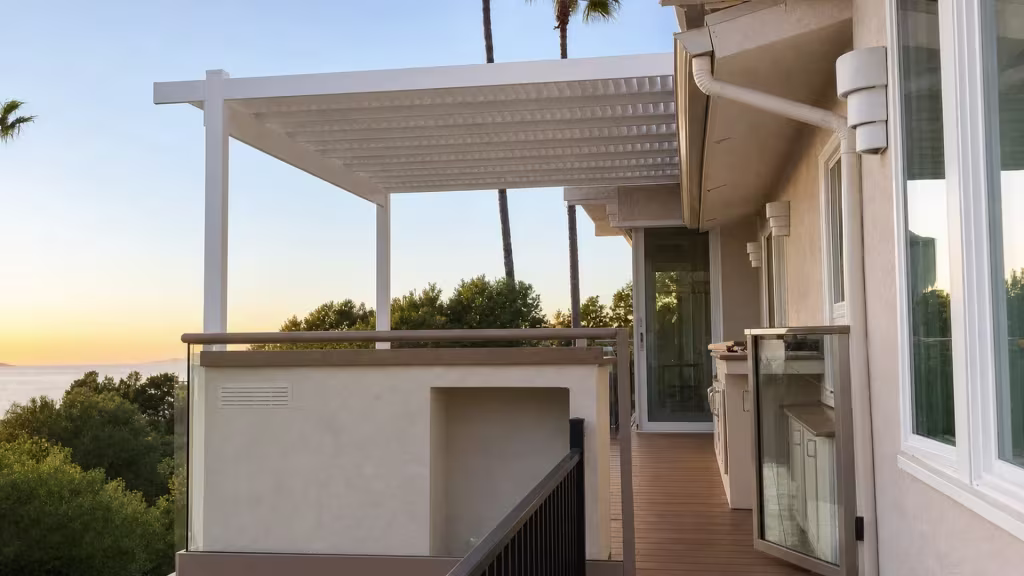 This louvered custom patio cover in White vinyl projects from a stucco building over a balcony with an ocean view