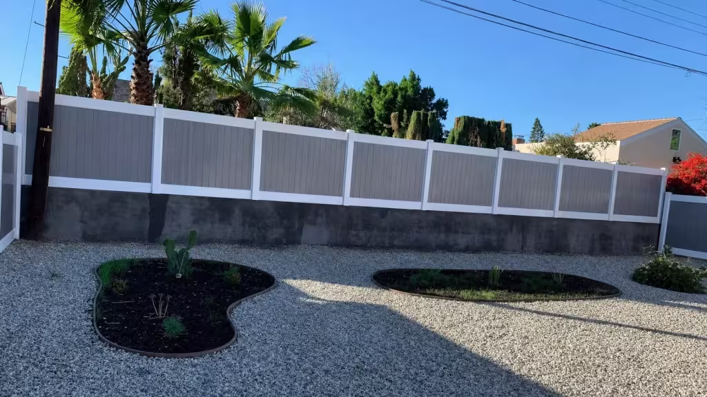 Tan custom vinyl wall topper fixed along a dark masonry wall bordering a gravel yard with curving planted beds