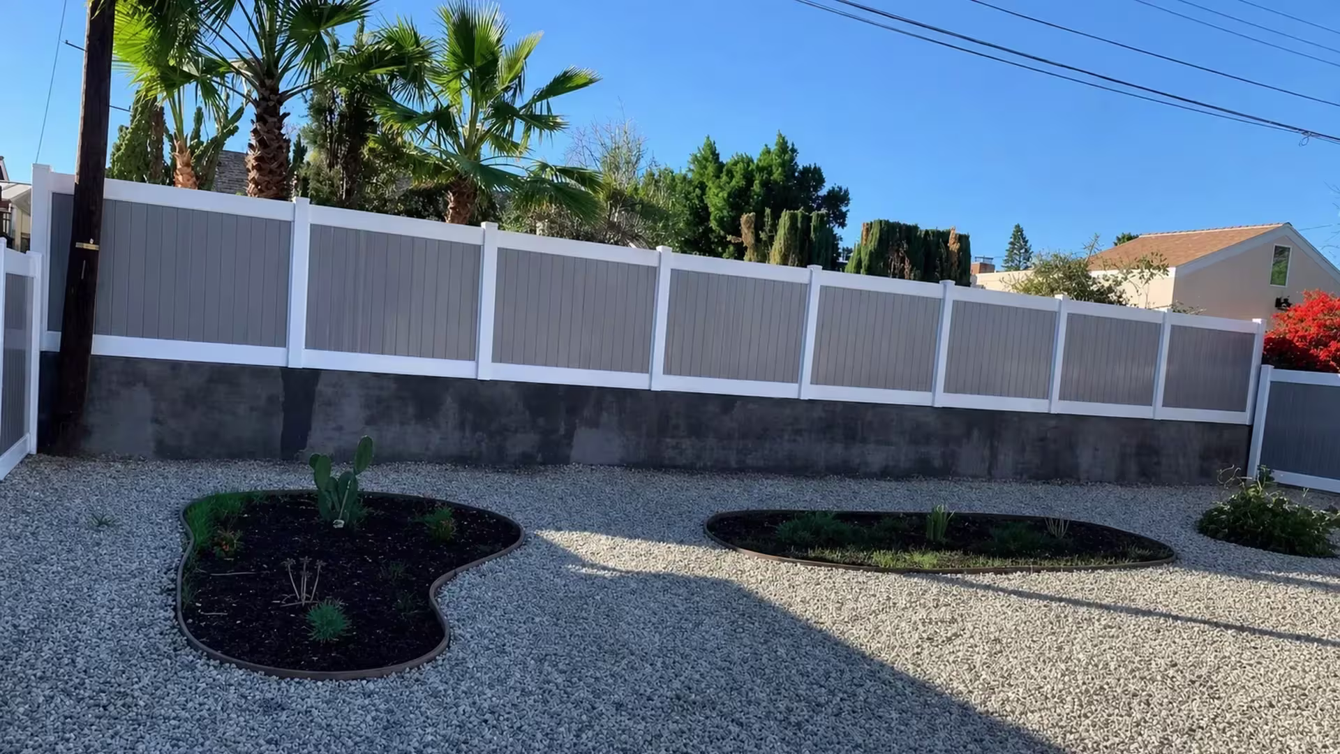 Tan custom vinyl wall topper fixed along a dark masonry wall bordering a gravel yard with curving planted beds