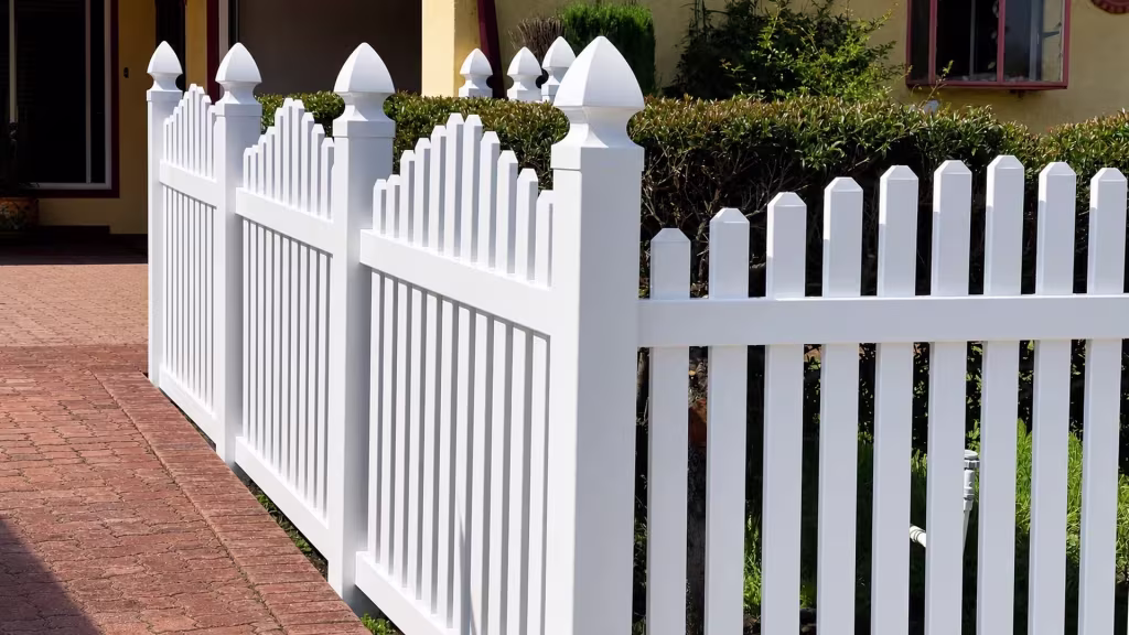 A brick-paved driveway curves beside a White vinyl dog-ear fence near the house