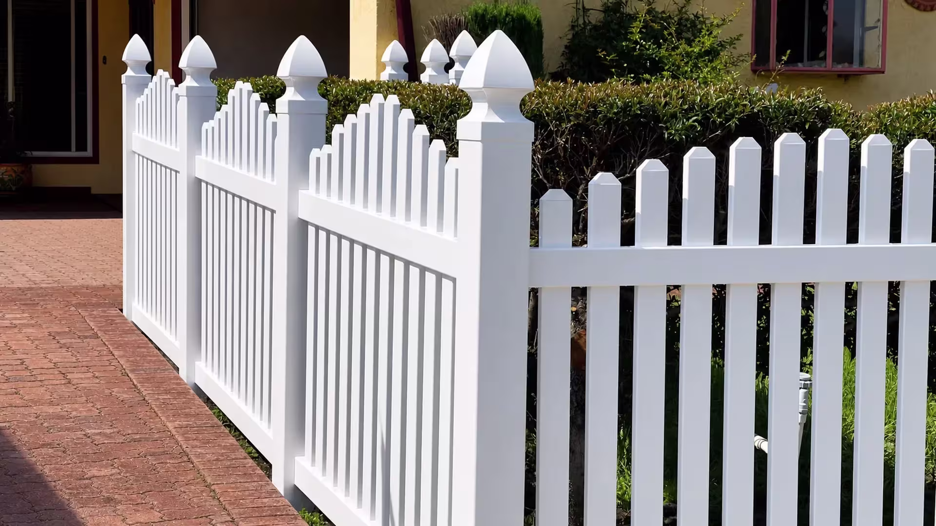 A brick-paved driveway curves beside a White vinyl dog-ear fence near the house