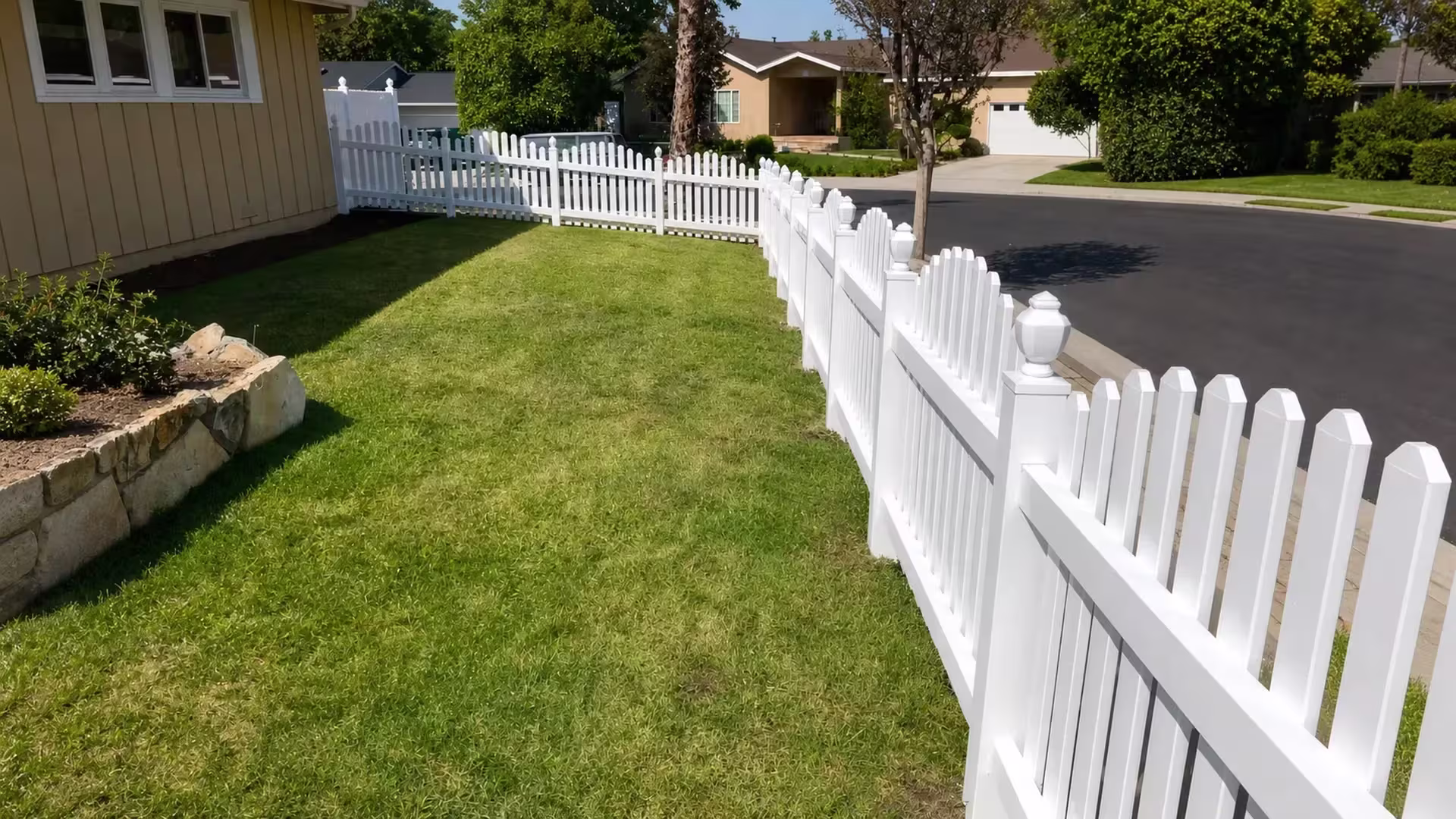 A corner lawn is bordered by a White vinyl dog-ear fence that curves past a stone planter