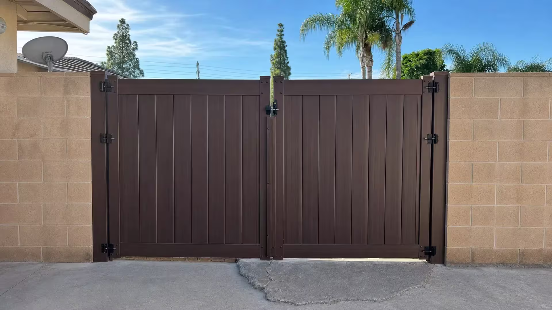 A Dark Walnut vinyl double-swing driveway gate closes the drive between tan concrete-block walls at a patched threshold