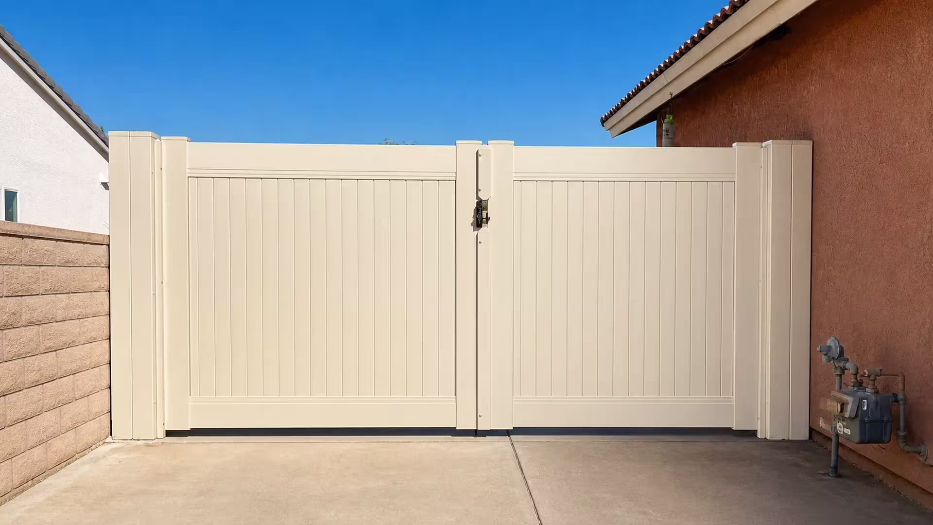 Tan vinyl driveway gate with unequal swinging leaves standing near a utility meter and tiled roofline