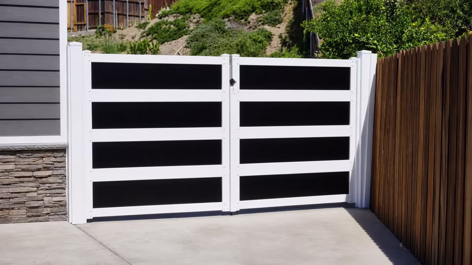 Glossy black horizontal infill bands mark this White vinyl driveway gate beside a stacked stone veneer wall