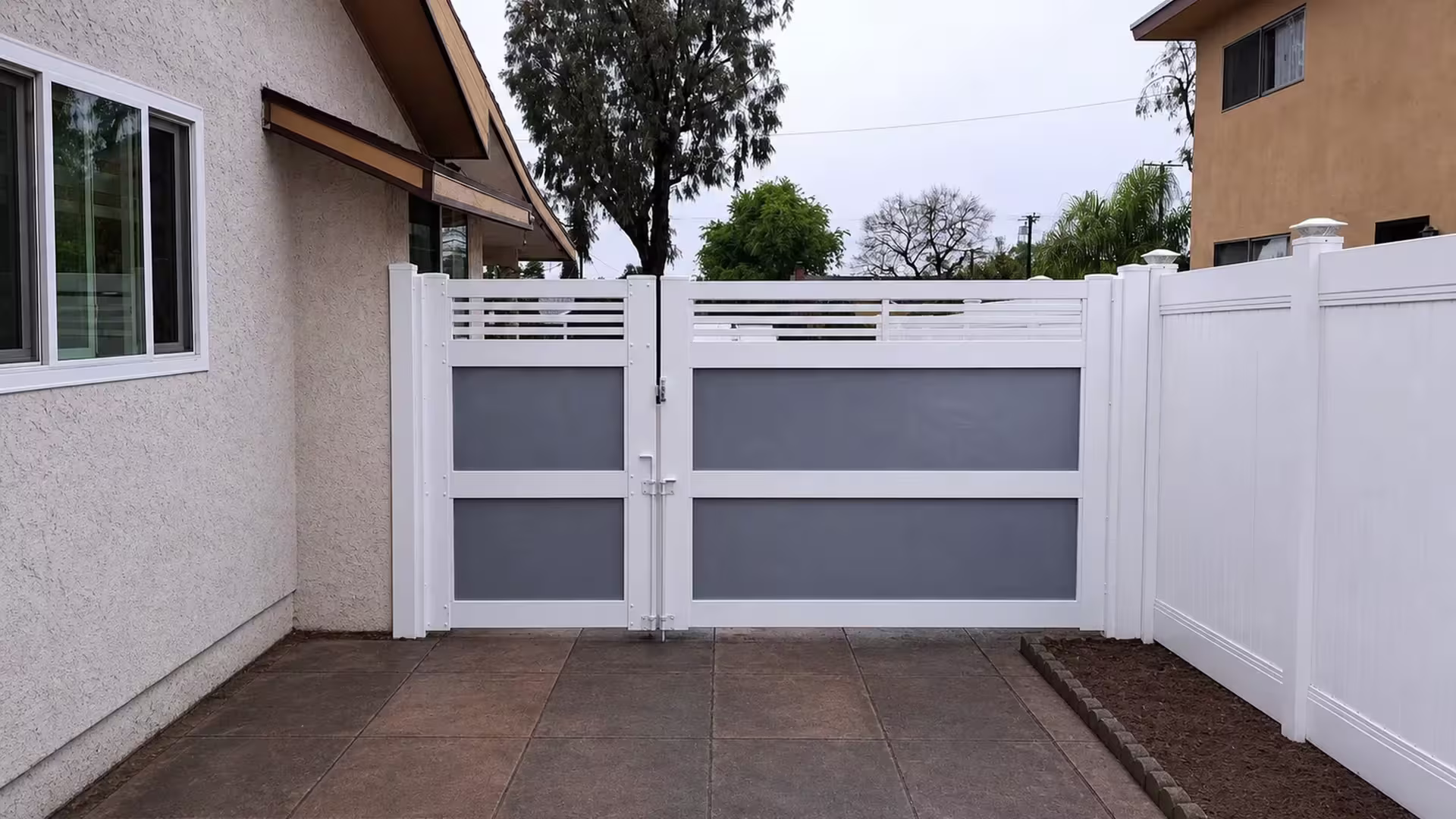 Stucco buildings flank this asymmetrical White vinyl double driveway gate with acrylic infill panels and palms nearby