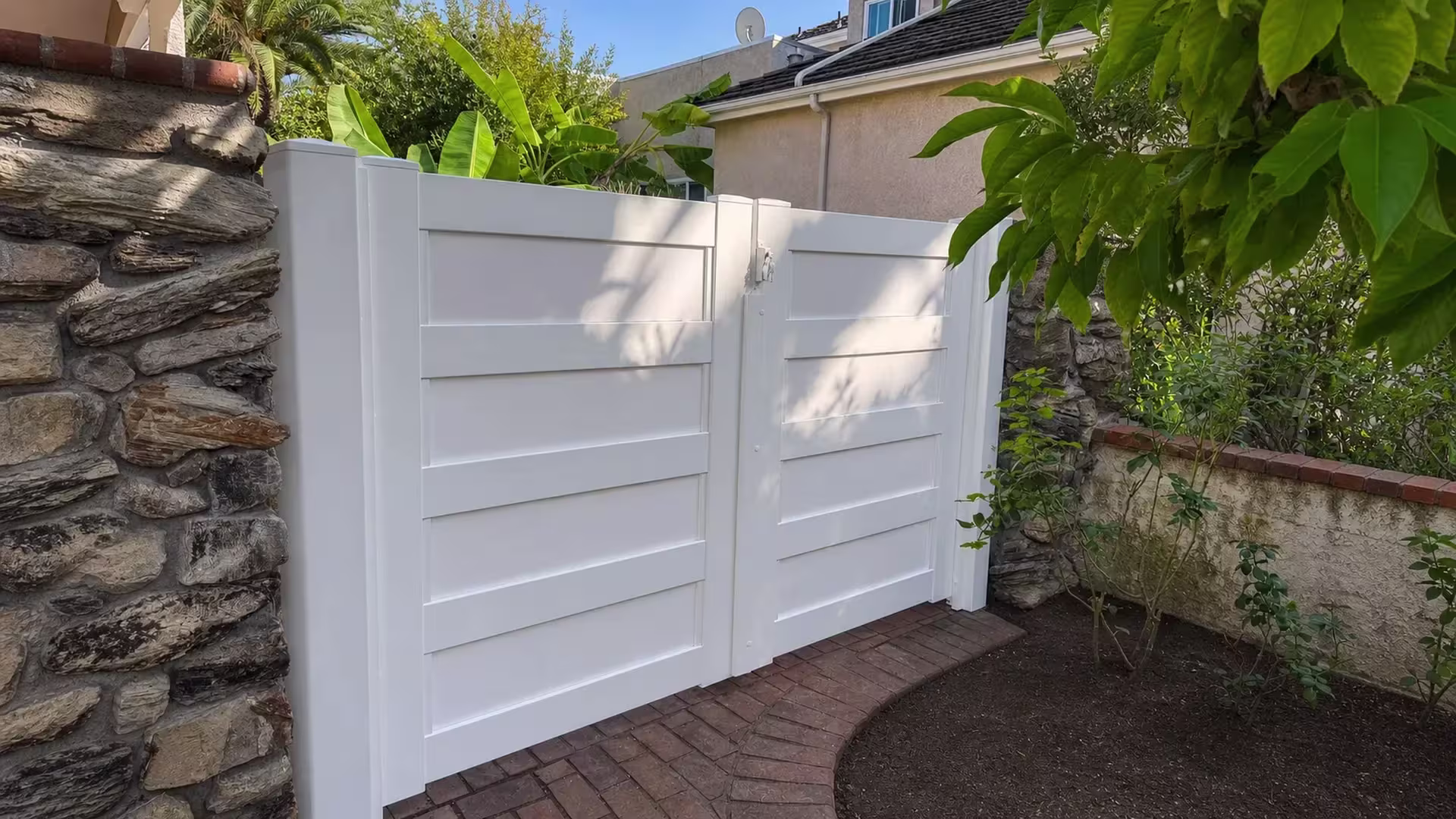 Stone boundary walls flank this unequal-leaf White vinyl driveway gate at the property edge