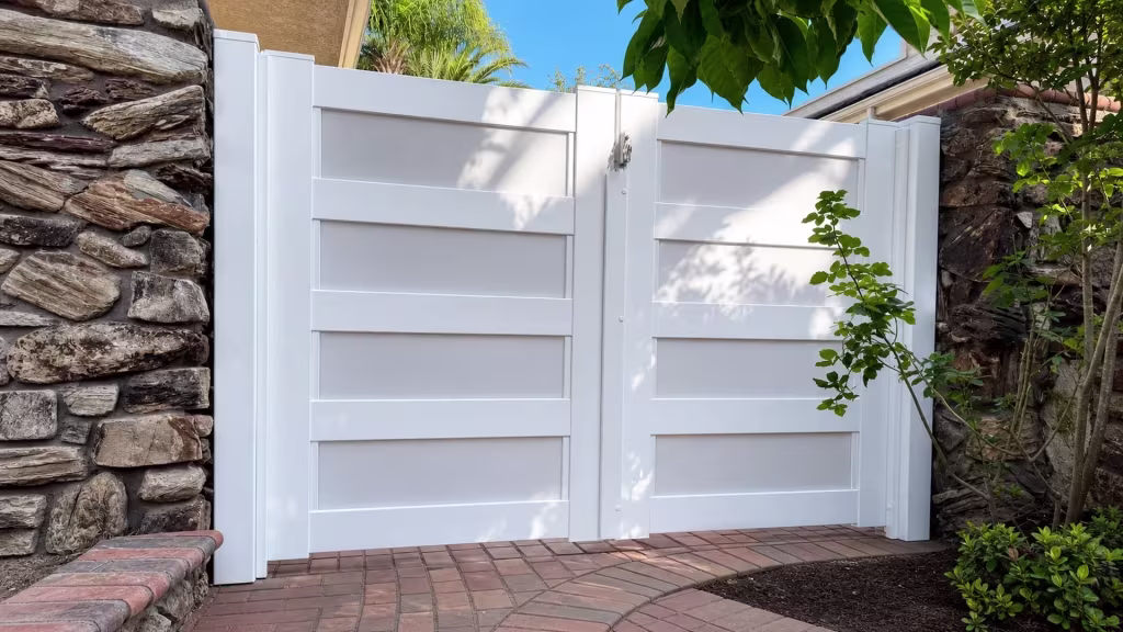Rough stone walls frame this White vinyl double driveway gate beside a brick-paved planting bed
