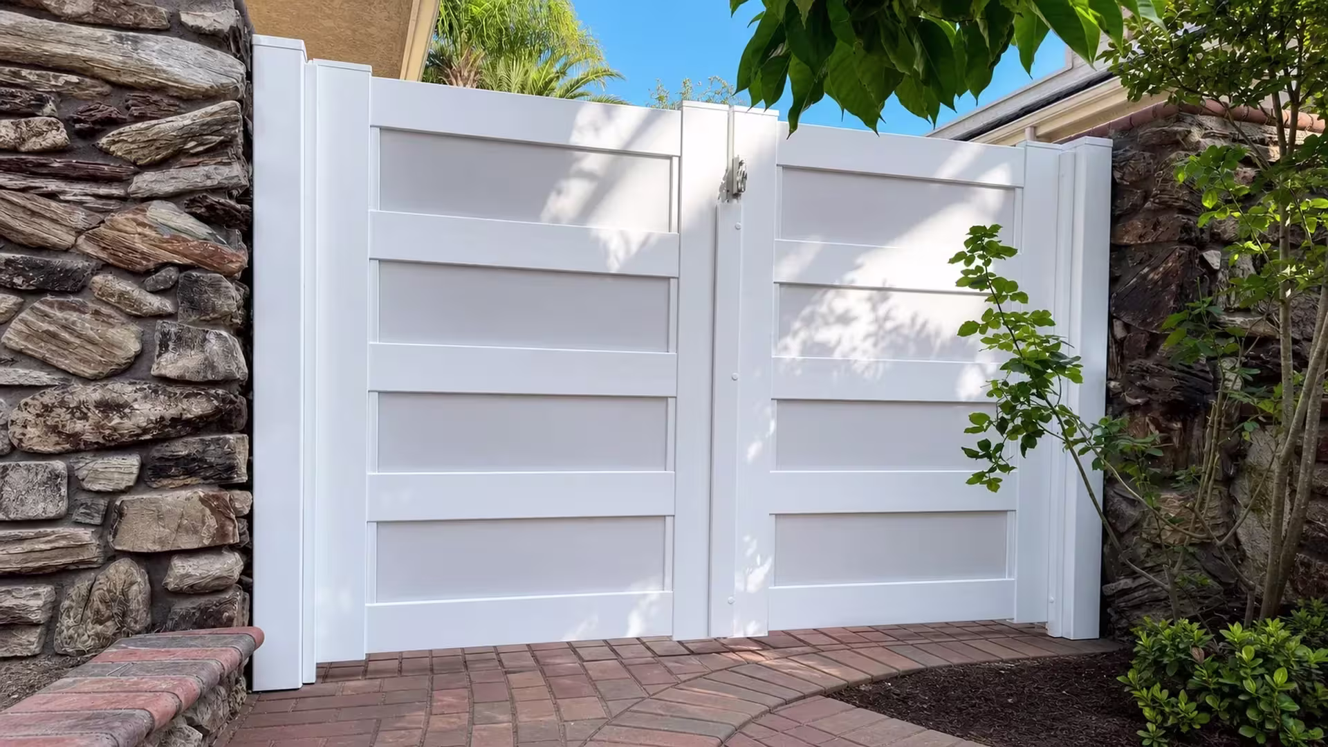 Rough stone walls frame this White vinyl double driveway gate beside a brick-paved planting bed