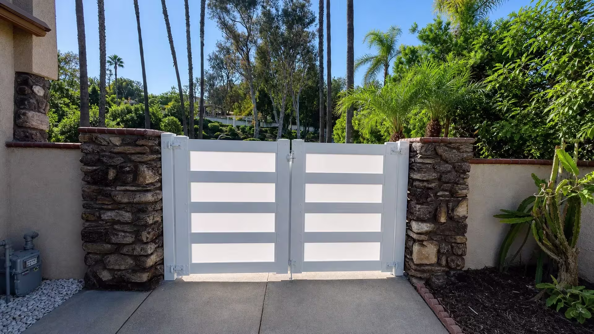 Rough-stone masonry columns anchor this White vinyl double driveway gate beside a planted brick-edged bed