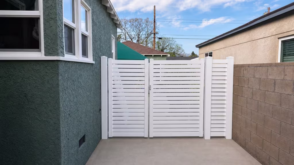 Between a green-gray stucco house and a tan block wall stands this White vinyl driveway gate with horizontal slats