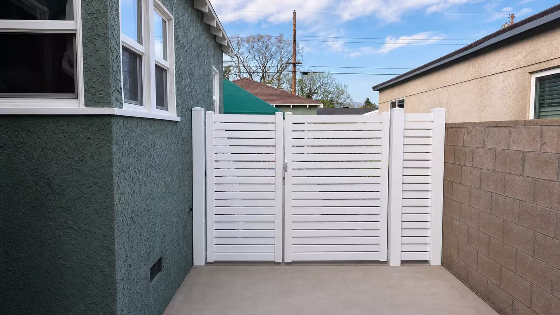 Between a green-gray stucco house and a tan block wall stands this White vinyl driveway gate with horizontal slats