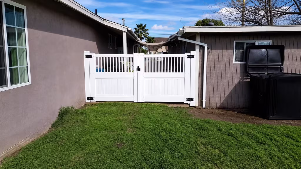 Two single-story buildings stand on either side of this White vinyl driveway gate, topped with an open picket band