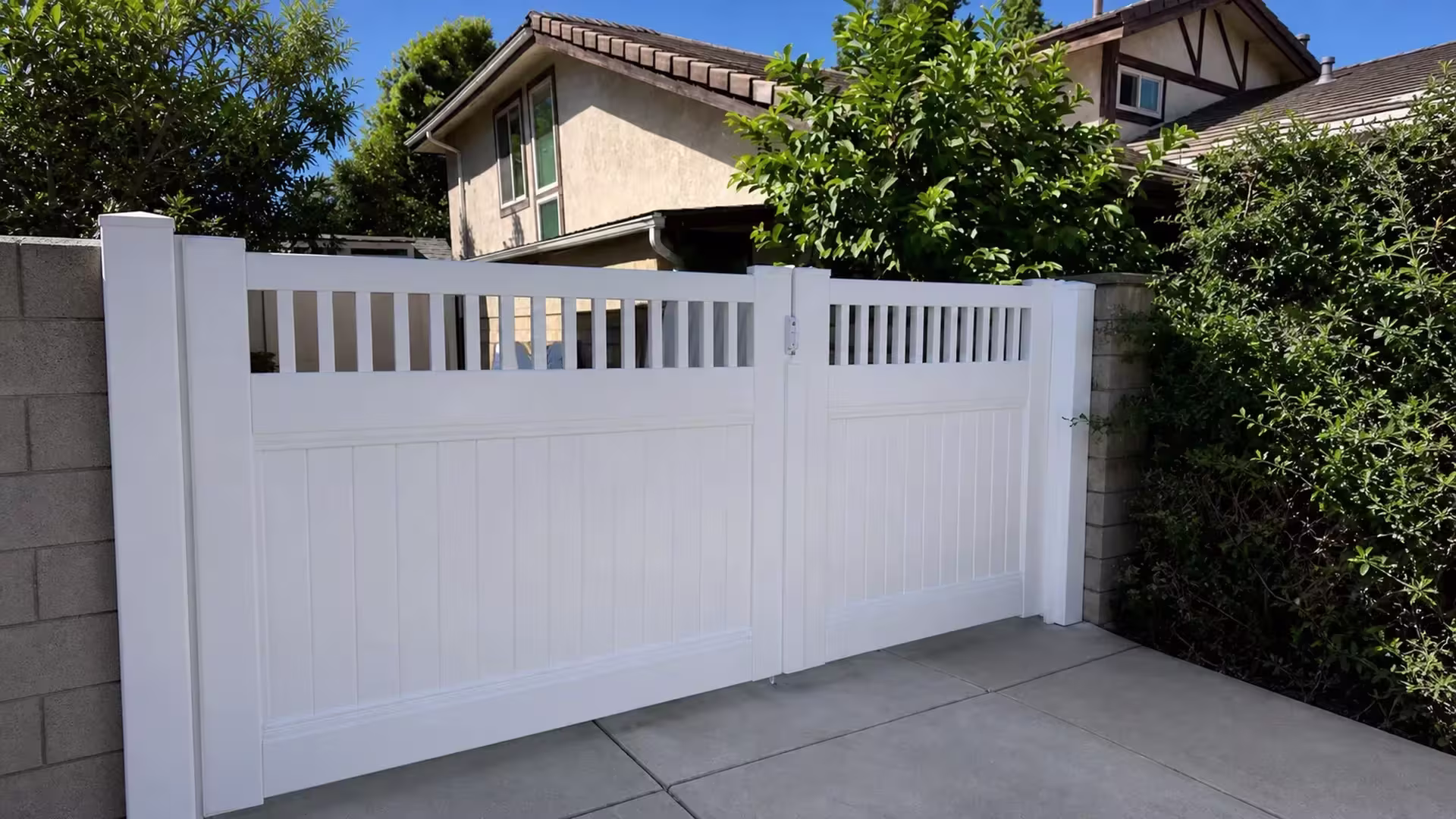 Tiled rooflines rise above this picket-top White vinyl double driveway gate beside a concrete block wall