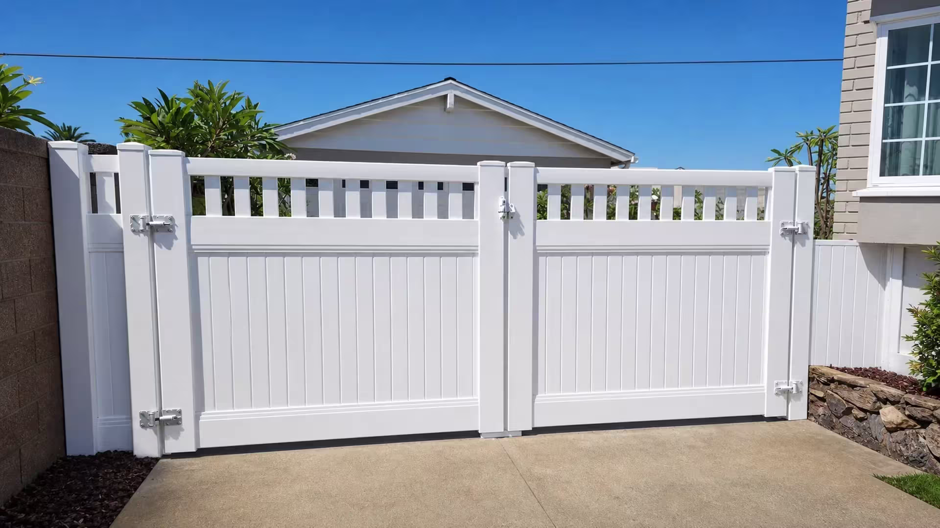 A detached gable roof rises behind this White vinyl driveway gate, its unequal leaves crowned with pickets