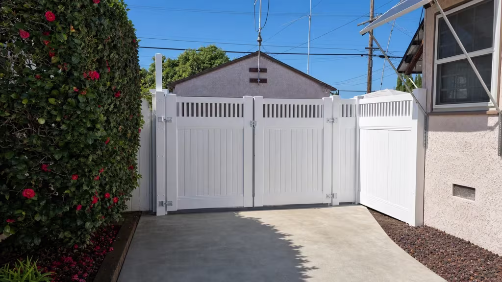 A flowering hedge and a metal awning flank this White vinyl driveway gate finished with open pickets