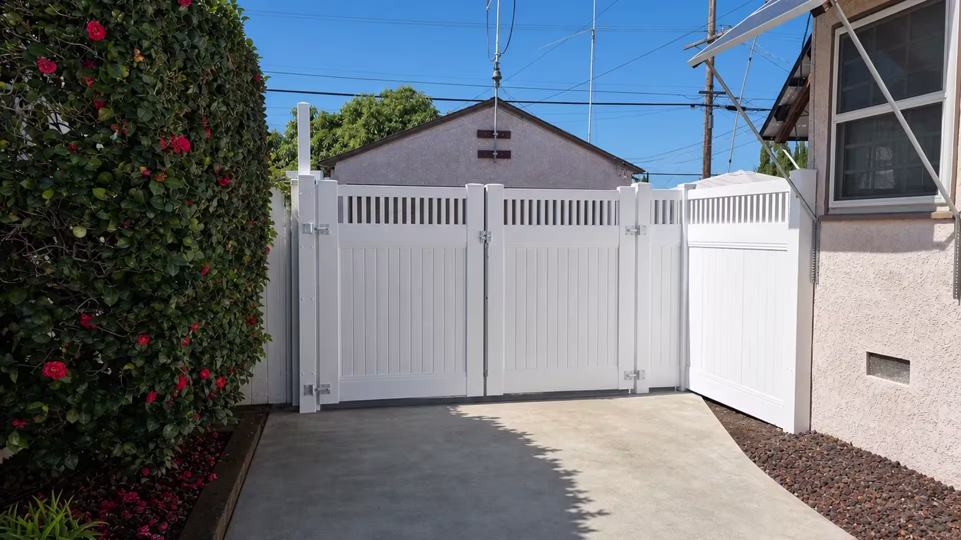 A flowering hedge and a metal awning flank this White vinyl driveway gate finished with open pickets