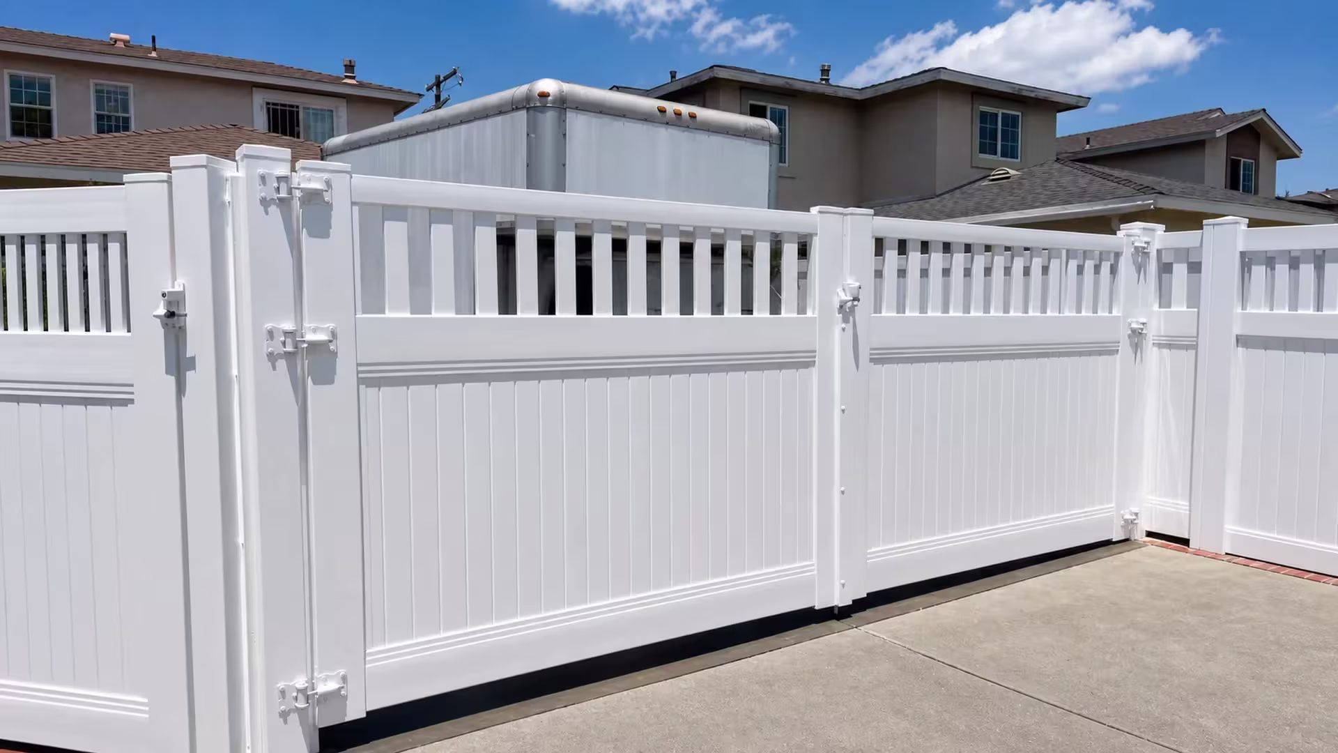 A light-gray enclosed trailer sits behind this White vinyl driveway gate, its lower boards faced with narrow pickets