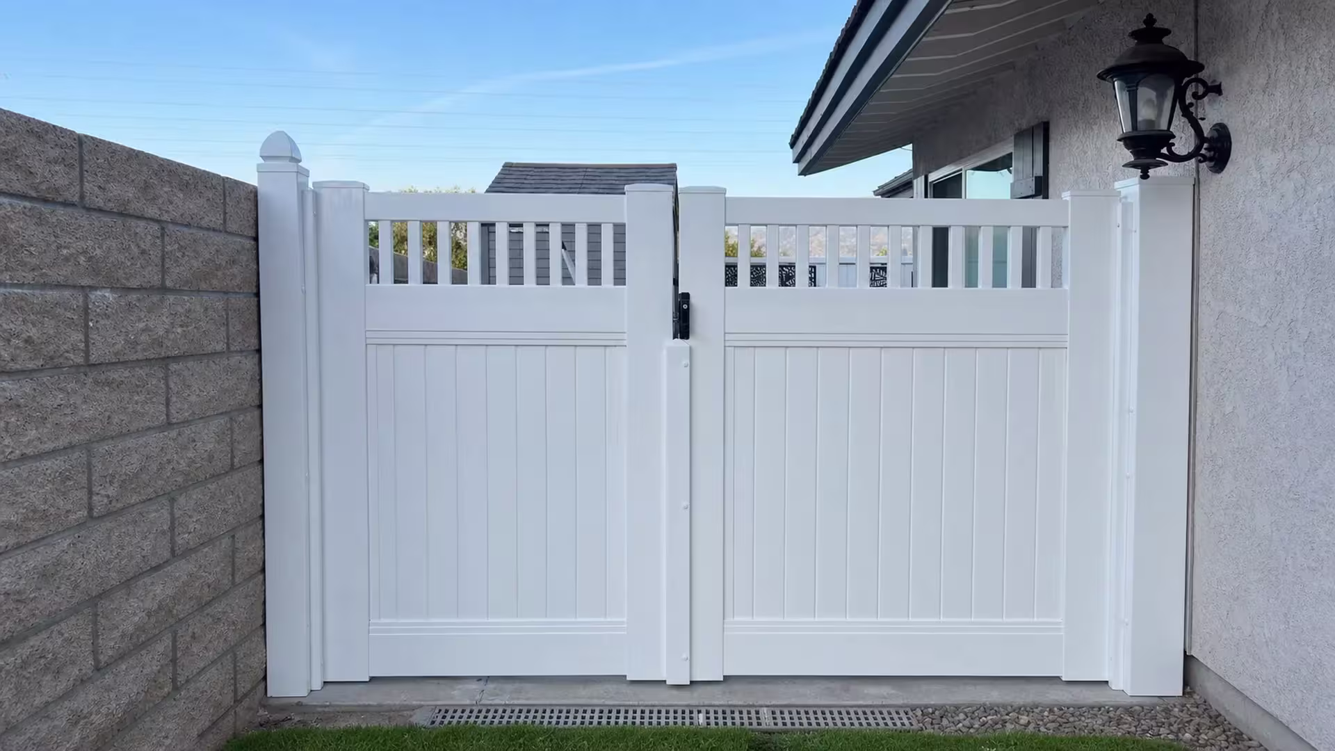 A black wall lantern hangs beside this White vinyl driveway gate, its picket top open above solid privacy boards