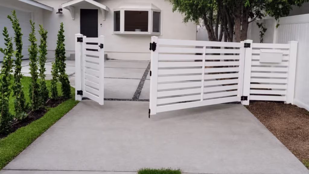 Standing open across the drive, this White vinyl ranch gate clears the entrance beside planting beds and trees