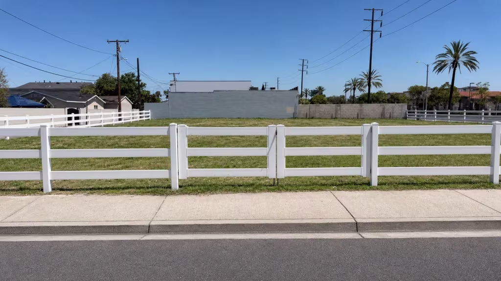 Diagonal metal braces reinforce this White vinyl three-rail ranch driveway gate, standing across from a vacant grassy lot
