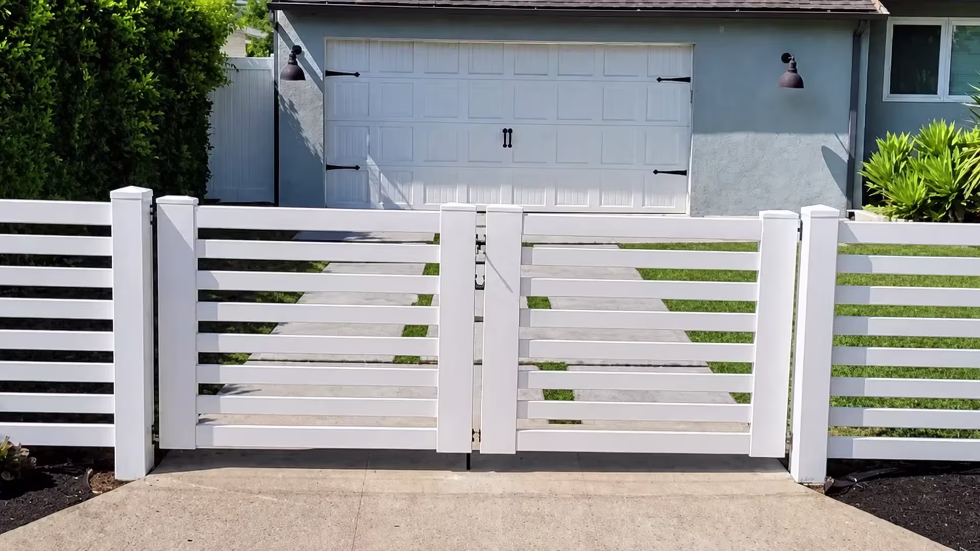 Yucca-like planting fills the bed beside this White vinyl horizontal-slat driveway gate in front of the gray home