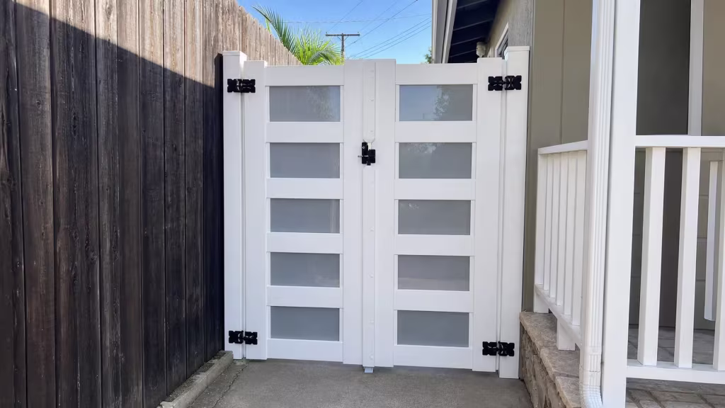 A dark weathered wood fence stands beside this White vinyl driveway gate, its leaves showing frosted infill panes