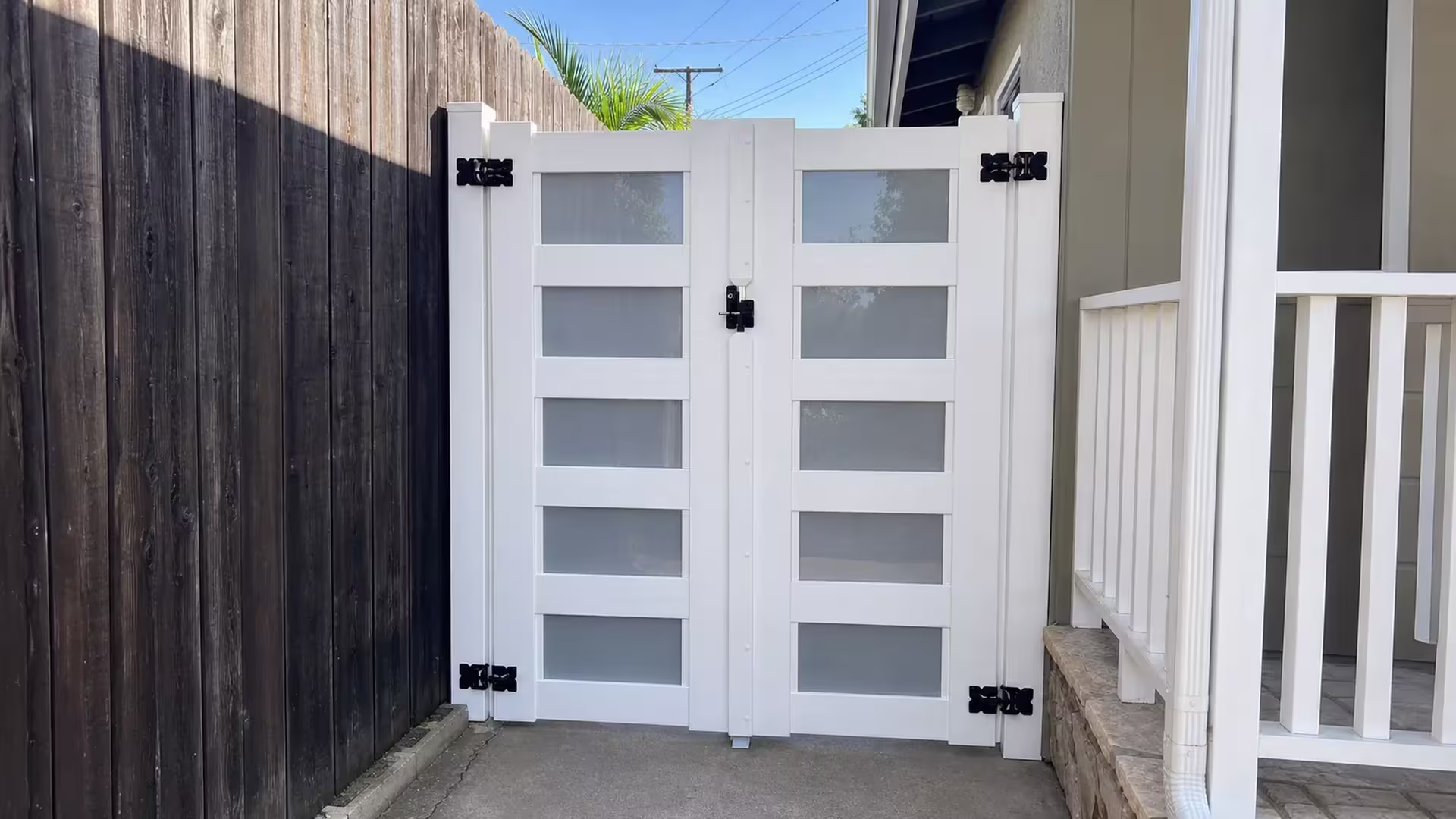 A dark weathered wood fence stands beside this White vinyl driveway gate, its leaves showing frosted infill panes