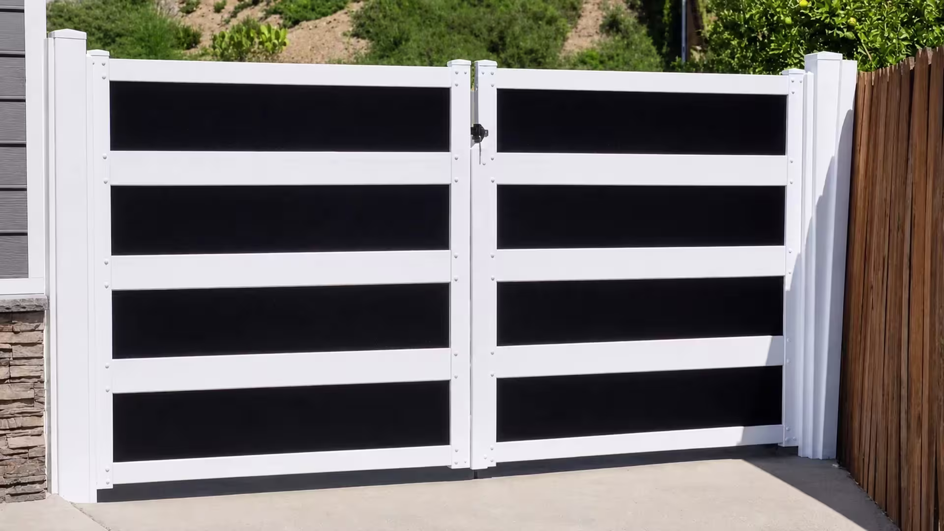 Gray stone veneer borders one side of this White vinyl driveway gate, a brown wood fence standing opposite it