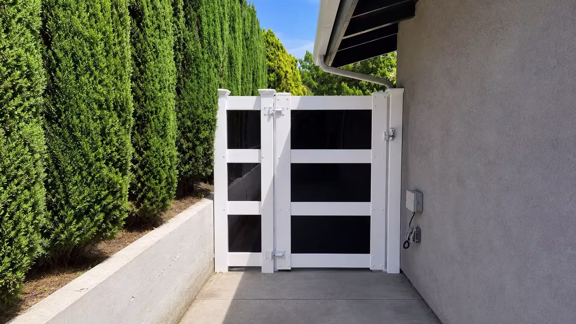 Tall columnar evergreens rise beside this White vinyl driveway gate in the narrow passage beside the stucco house