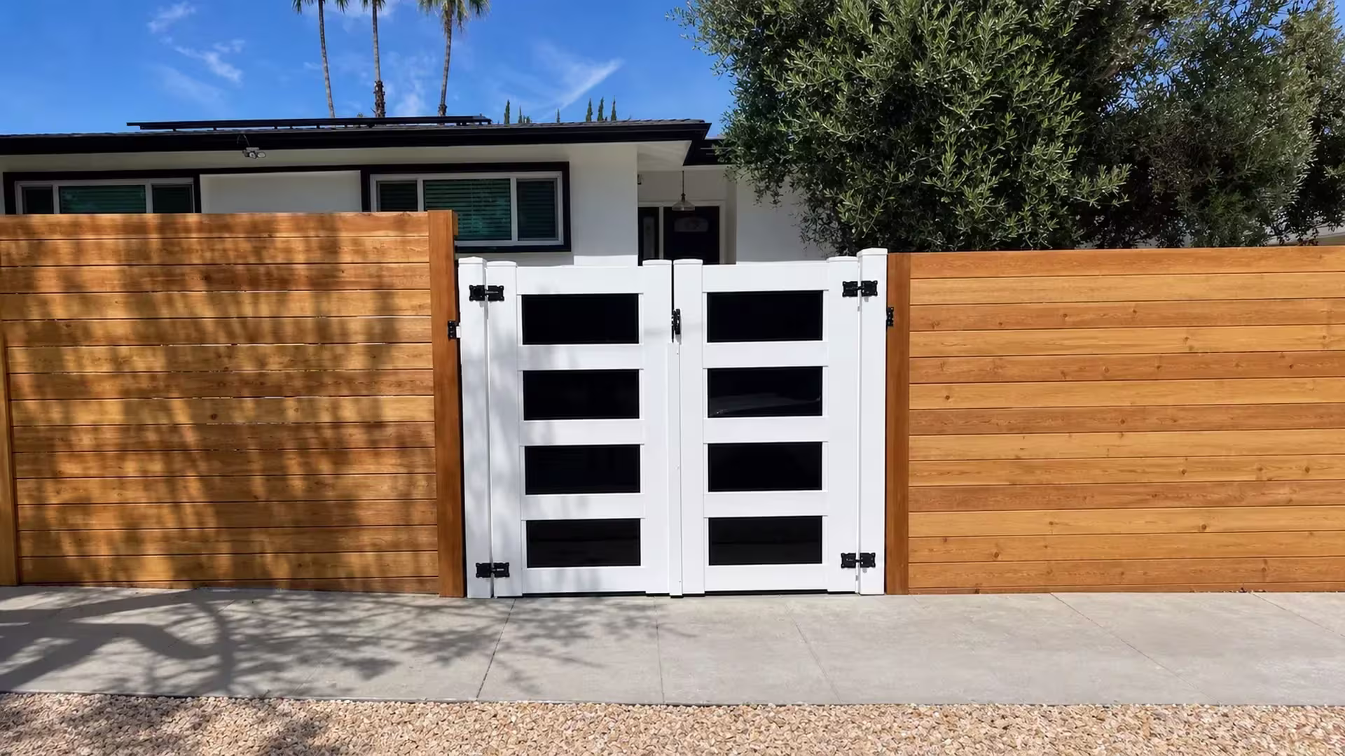 Warm horizontal wood fencing flanks both sides of this White vinyl driveway gate, centered on the concrete walkway