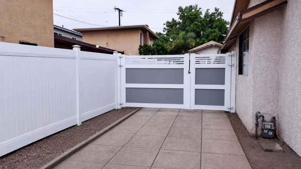 A gas meter and pipes sit beside this White vinyl driveway gate, its left leaf noticeably wider than the right