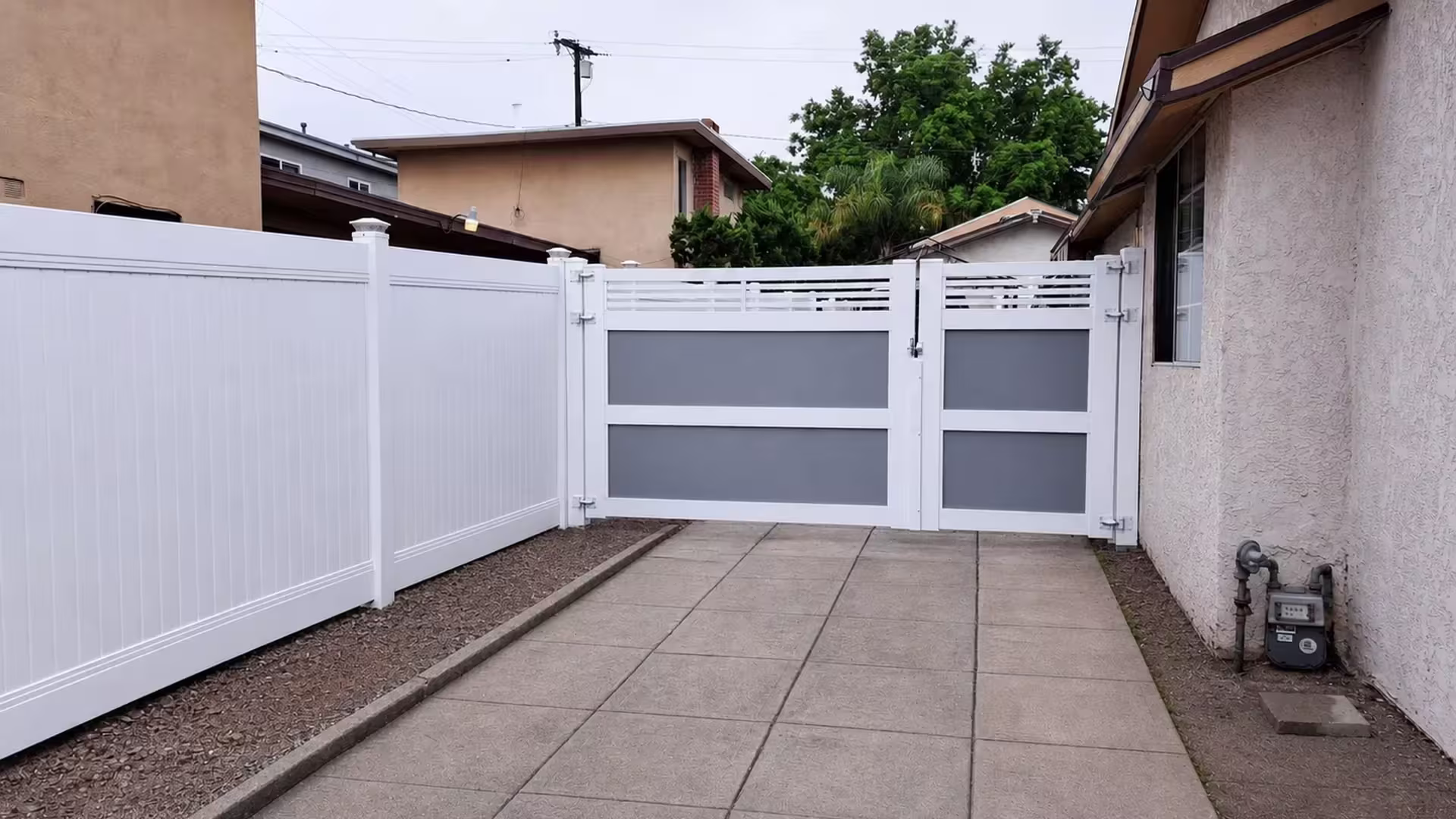 A gas meter and pipes sit beside this White vinyl driveway gate, its left leaf noticeably wider than the right